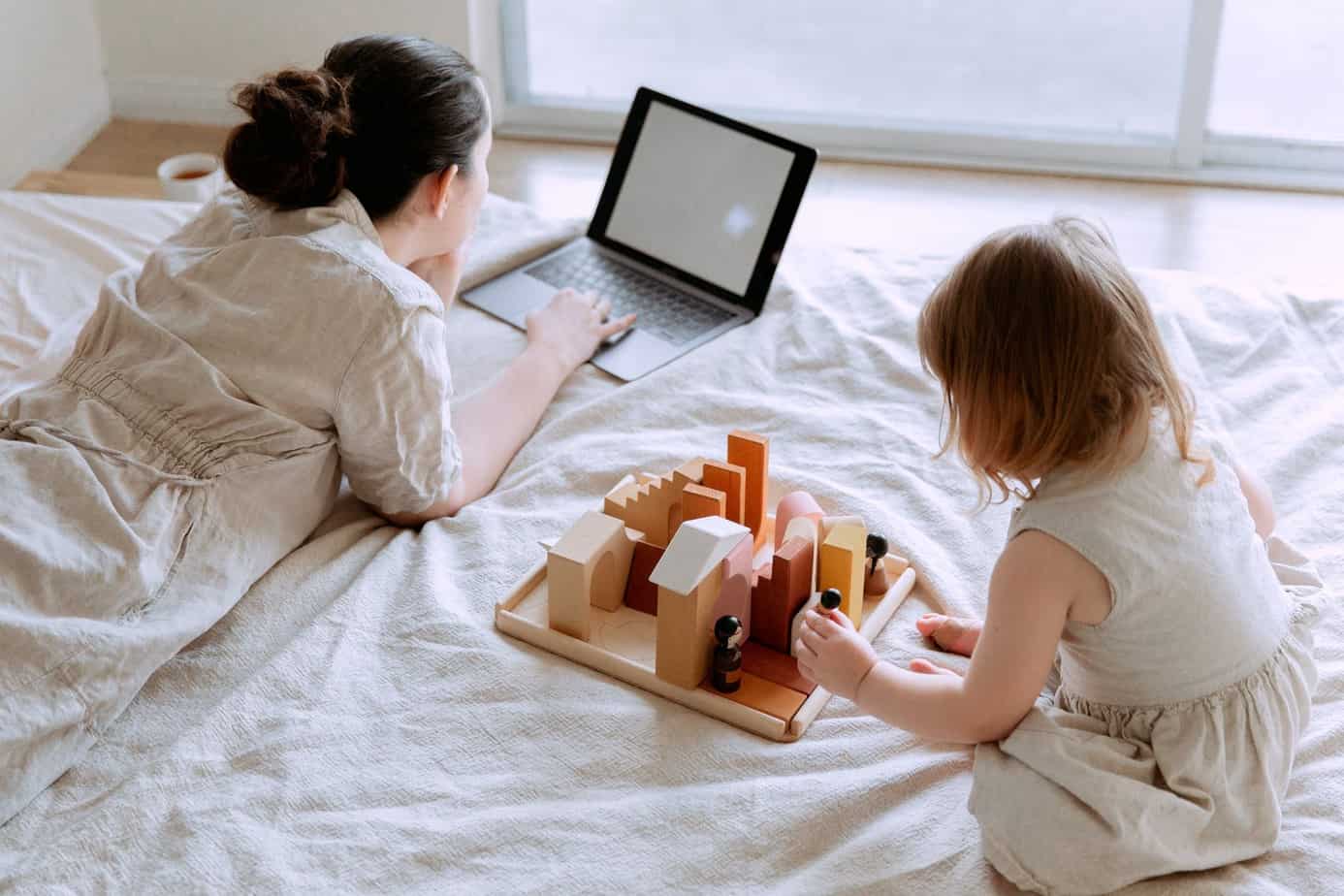 young woman lying on bed and using laptop while daughter playing with wooden constructor